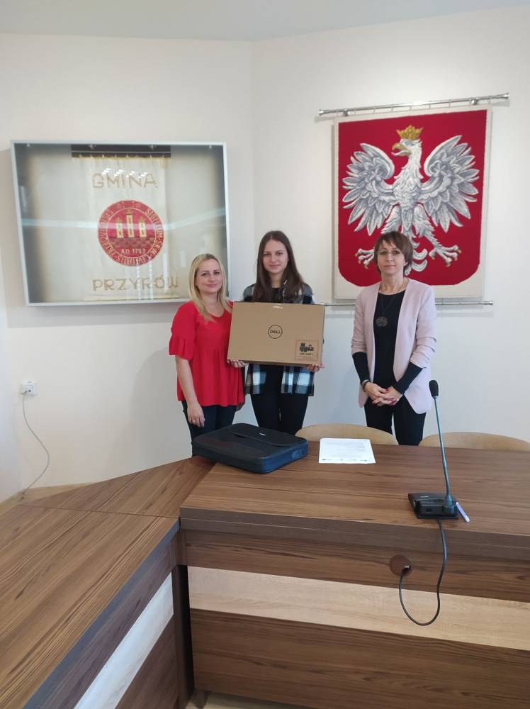 : Dziewczynka trzymająca laptopa wraz z opiekunem oraz Pani Dorota Wojciechowska - Sekretarz Gminy.